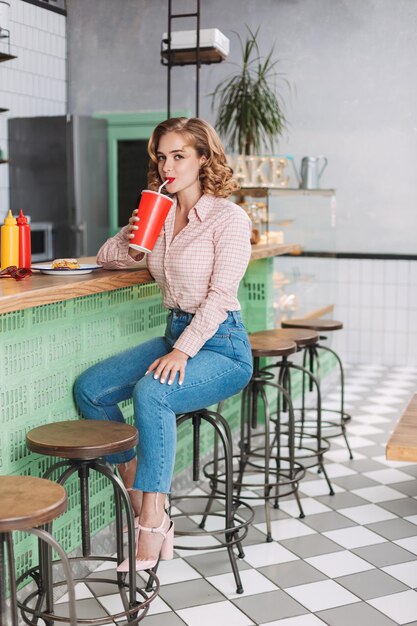 Hübsche Dame in Hemd und Jeans, die an der Bartheke sitzt und Sodawasser trinkt, während sie glücklich in die Kamera im Café blickt