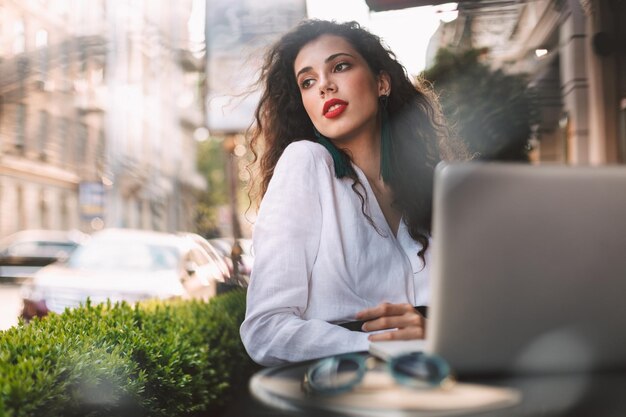 Hübsche Dame mit dunklem lockigem Haar in weißem Kostüm, die mit Laptop am Tisch sitzt und verträumt zur Seite schaut, während sie Zeit im Café auf der Straße mit Blick auf die Stadt im Hintergrund verbringt