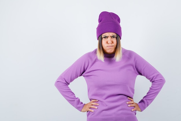 Kostenloses Foto hübsche dame mit händen an der taille, blasenden wangen in pullover, mütze und boshaftem aussehen, vorderansicht.