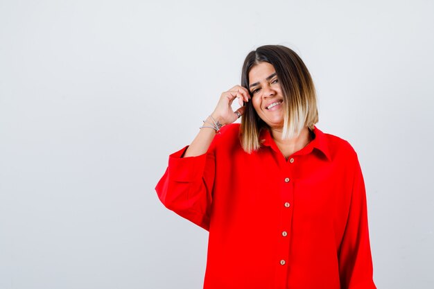 Hübsche Dame posiert beim Berühren der Haare in roter Bluse und sieht fröhlich aus, Vorderansicht.