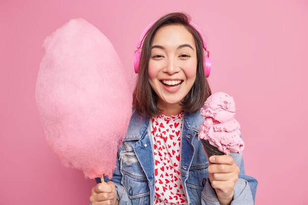 Hübsche dunkelhaarige junge Frau mit positivem Ausdruck hält gefrorenes Eis und Zuckerwatte hört Audiospur über Kopfhörer in modischer Kleidung isoliert über rosa Wand pink