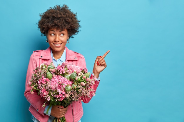 Hübsche dunkelhäutige hübsche junge Frau mit Afro-Haaren zeigt an, dass auf dem Kopierraum Bouquet Empfehlungen gibt, die über blauem Studiohintergrund isoliert werden.