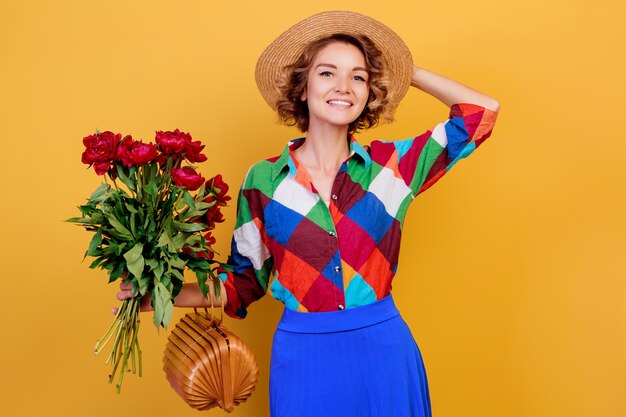 Hübsche europäische Frau im blauen Kleid, das Blumenstrauß über gelbem Hintergrund hält. Strohhut. Sommerstimmung.