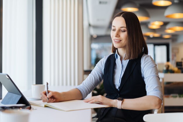 Hübsche Frau, die Arbeitszeitplan schreibt, der in Notizbuch schreibt, während sie am Arbeitsplatz mit Tablette sitzt.