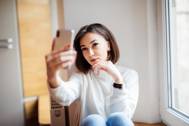 Hübsche Frau, die auf Fensterbank im weißen Pullover-Selfie auf ihrem Telefon sitzt