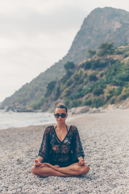 Hübsche Frau, die im Strand im schwarzen Strandkleid während des Tages sitzt und meditiert
