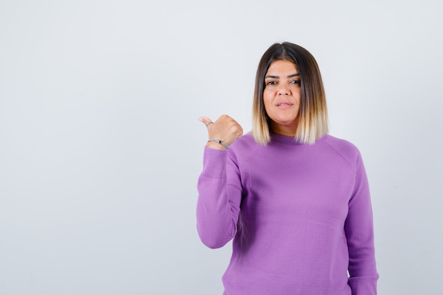 Hübsche Frau, die mit dem Daumen im lila Pullover zur Seite zeigt und selbstbewusst aussieht, Vorderansicht.