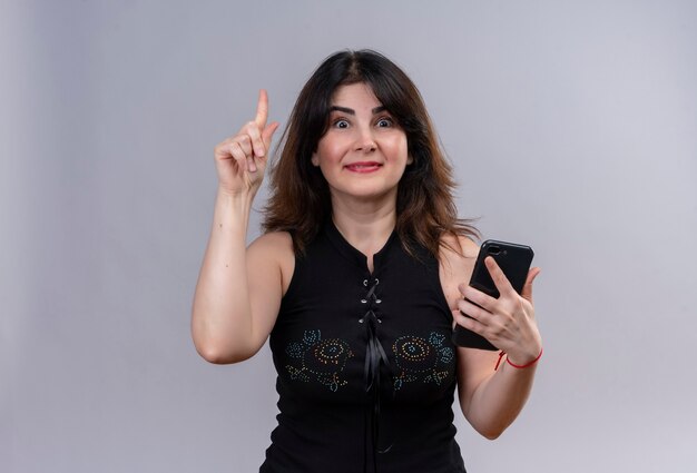 Hübsche Frau, die schwarze Bluse trägt, die glückliche Zeigefinger-Gründungslösung am Telefon tut