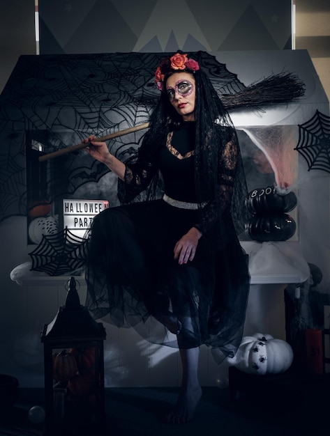 Kostenloses Foto hübsche frau feiert halloween in schwarzem kleid und ungewöhnlichem make-up, während sie besen hält.