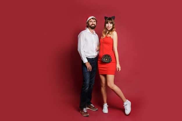 Hübsche Frau im Katzenohren-Maskeradenhut und im roten kurzen Kleid mit ihrem Freund, der auf Rot aufwirft. Neujahrsparty. In voller Länge .