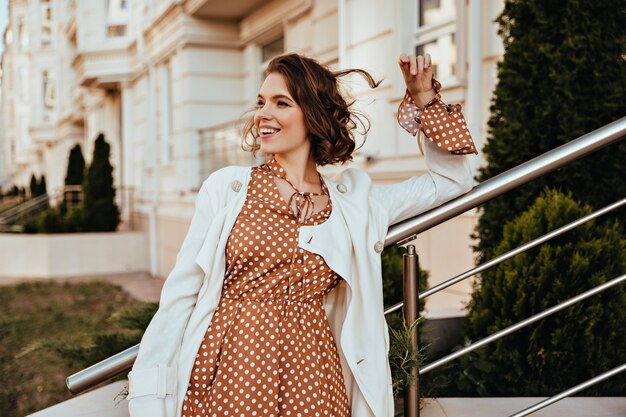 Hübsche Frau im langen braunen Kleid, das auf der Straße lächelt. Außenaufnahme des emotionalen weiblichen Modells mit elegantem Make-up.