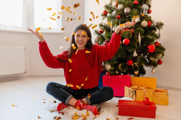 Hübsche Frau im roten Pullover sitzt zu Hause am Weihnachtsbaum und wirft goldene Konfetti, umgeben von Geschenken und Geschenkboxen