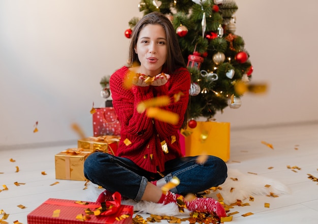 Hübsche Frau im roten Pullover sitzt zu Hause am Weihnachtsbaum und wirft goldene Konfetti, umgeben von Geschenken und Geschenkboxen