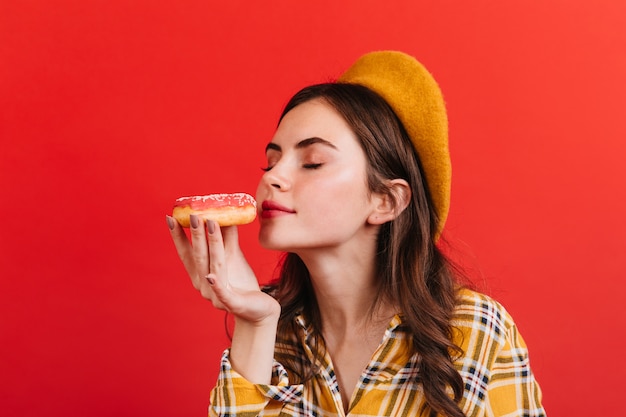 Kostenloses Foto hübsche frau in baskenmütze schnüffelt duftenden donut. porträt des mädchens mit dem lockigen haar auf der isolierten roten wand.