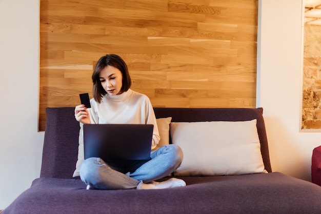 Hübsche Frau in den blauen Jeans und im weißen T-Shirt arbeitet am Laptop, der auf dunklem Bett vor Holzwand zu Hause sitzt