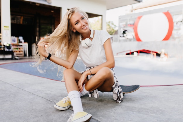 Hübsche Frau in den gelben Schuhen, die auf Longboard mit gekreuzten Beinen sitzen. Foto im Freien des atemberaubenden gebräunten weiblichen Modells, das Spaß im Skatepark hat und mit ihren Haaren spielt.