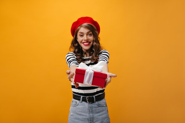 Hübsche Frau in der roten Baskenmütze demonstriert Mädchenbox. Glückliches Mädchen mit welligem Haar in den modernen Kleidern, die auf orange Hintergrund lächeln.