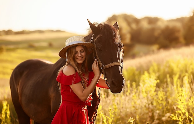 Hübsche Frau in einem Heuhut und in einem roten Kleid steht mit einem Pferd auf dem grünen Feld