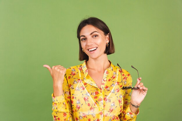 Hübsche Frau in gelbem Sommerkleid und Sonnenbrille, grünes Studio, aufgeregter fröhlicher, fröhlicher Zeigefinger links