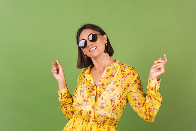 Hübsche Frau in gelbem Sommerkleid und Sonnenbrille, grünes Studio, glückliche positive fröhliche freudige Emotionen