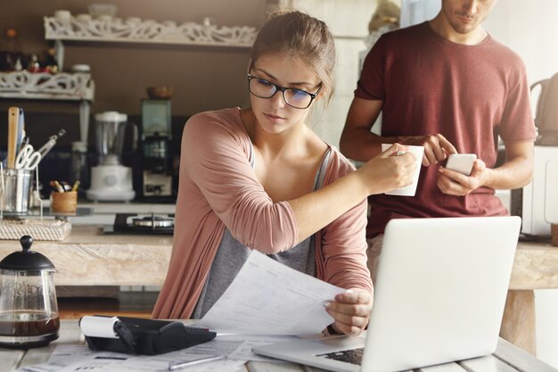 Hübsche Frau in Gläsern, die ernst aussehen, während sie Hausbudget tun, am Tisch in der Küche sitzen, Rechnung in ihrer Hand studieren und Tasse Tasse an ihren Ehemann weitergeben