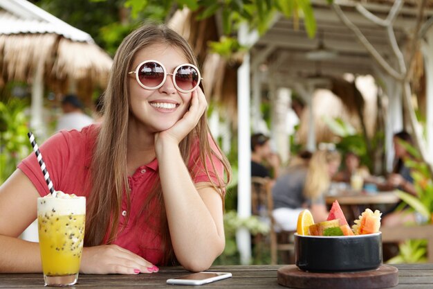 Hübsche Frau in runder Sonnenbrille, die lang erwarteten Urlaub im tropischen Land genießt, Fruchtcocktail hat, ihren Ellbogen auf Kaffeetisch mit Handy darauf ruht, fröhlichen und entspannten Ausdruck hat