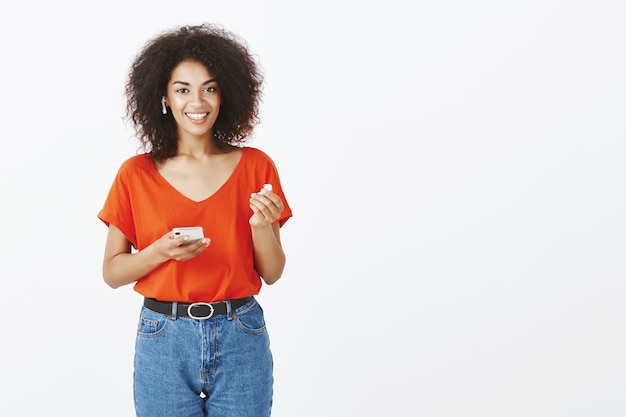 Hübsche Frau mit Afro-Frisur, die mit ihrem Smartphone und Ohrhörern im Studio aufwirft