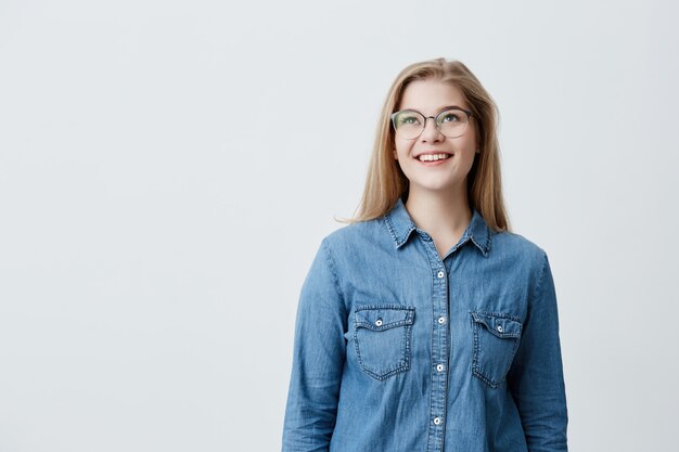 Hübsche Frau mit blasser Haut und breitem glücklichem Lächeln, das große runde Brillen trägt, nach oben schaut, gute positive Nachrichten über ihre Beförderung bei der Arbeit genießt und gegen grauen leeren Hintergrund aufwirft