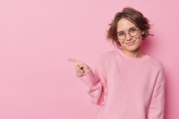 Hübsche Frau mit trendiger Frisur, fröhlicher Ausdruck weist auf Werbebanner hin, oder Rabatt trägt runde Brille und Pullover isoliert über rosa Hintergrund, stellt neues Produkt vor