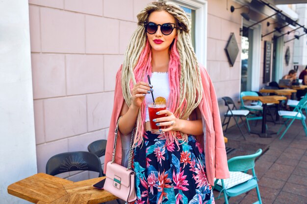 Hübsche Frau mit ungewöhnlicher Dreads-Frisur, die auf der Straße posiert, rosa Blumen femininen eleganten Blick trägt und frische Limonade hält