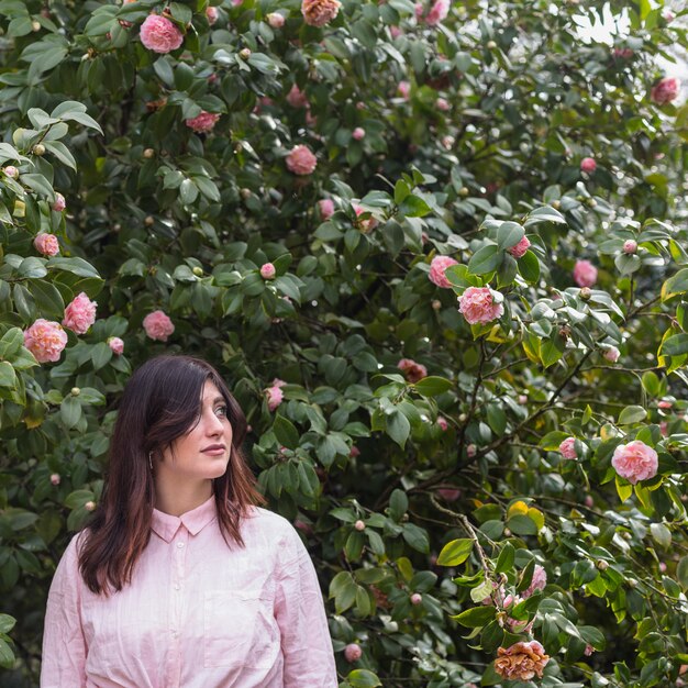 Hübsche Frau nahe den rosa Blumen, die auf grünen Zweigen wachsen