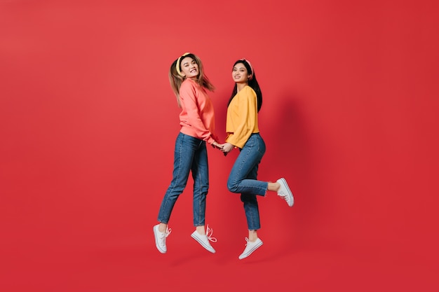 Hübsche Frauen in stilvollen Sweatshirts und Jeans springen auf rote Wand