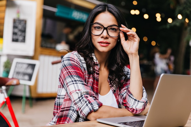 Hübsche Freiberuflerin trägt eine trendige Brille, die auf Unschärfestadt posiert. Elegantes schwarzhaariges Mädchen mit Laptop an einem guten Tag.