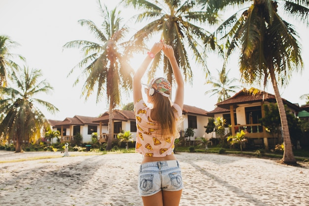 Hübsche Hipster-Frau, die auf Strandtanz geht, der Musik auf Kopfhörern in stilvollem buntem Outfit an sonnigen tropischen Sommerferien hört, die Zubehörkappen-Sonnenbrille tragen und Spaß haben