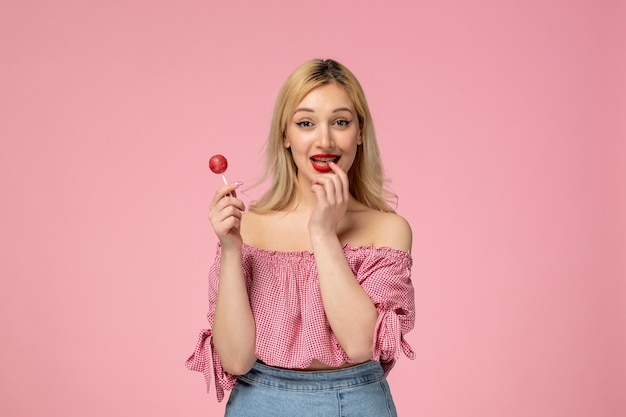 Hübsche junge Dame des netten Mädchens mit rotem Lippenstift in der rosafarbenen Bluse, die den Finger hält, der eine Süßigkeit hält