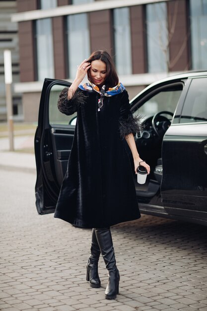 Hübsche junge Dame, die einen modischen schwarzen Mantel trägt, während sie Kaffee hält und in der Nähe des Autos steht. Konzept der Modestadt