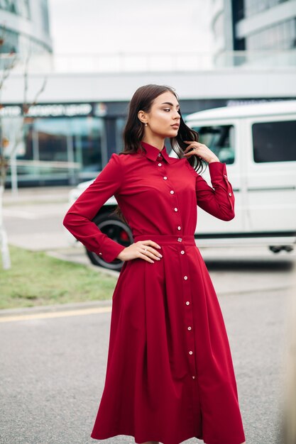 Hübsche junge Dame, die rotes Kleid trägt, während auf der Straße mit Auto und Gebäude auf dem Hintergrund aufwirft. Stadtlebensstil