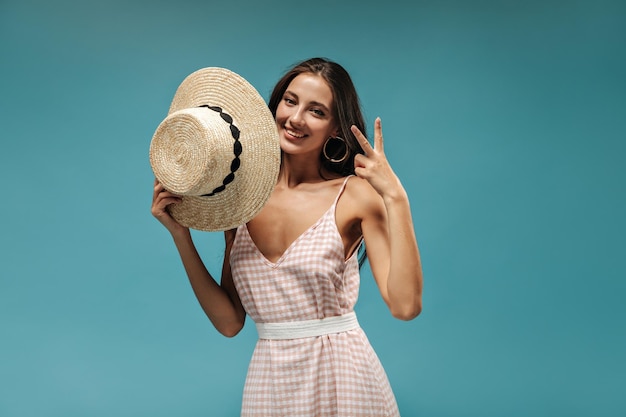 Hübsche junge Dame in sommerlicher rosa Kleidung lächelt, zeigt Peace-Zeichen und hält modernen Hut auf isoliertem blauem Hintergrund