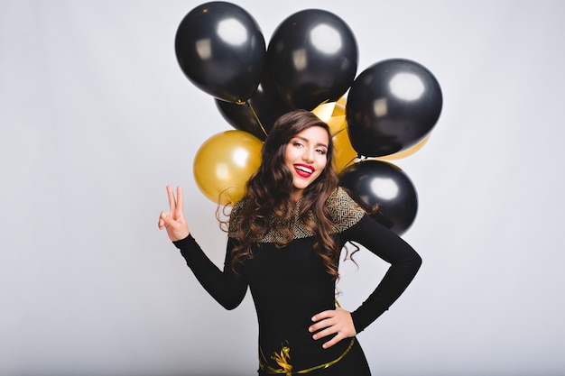 Kostenloses Foto hübsche junge frau auf weißem raum halten goldene und schwarze luftballons. erstaunliches mädchen mit langen lockigen haaren, im schwarzen eleganten kleid