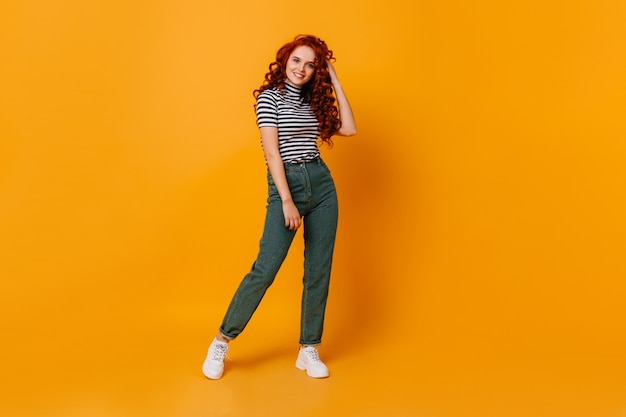 Hübsche junge Frau berührt ihr lockiges rotes Haar und lächelt. Porträt des Mädchens in der Jeans und im Top im orangefarbenen Studio.