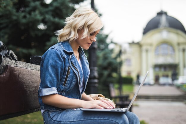 Hübsche junge Frau, die auf der Bank sitzt und Telefon und Laptop im Herbstmorgen der Stadt benutzt