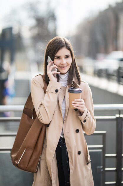 Hübsche junge Frau, die die Straße geht, die Kaffeetasse und Telefon hält