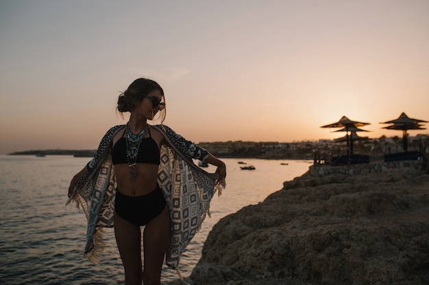 Hübsche junge Frau, die Sonnenuntergang am Strand mit Felsen genießt und Urlaub hat. Zur Seite schauen. Tragen Sie eine stilvolle Sonnenbrille, einen schwarzen modischen Badeanzug, einen Bikini, eine Strickjacke und einen Umhang mit Ornamenten.