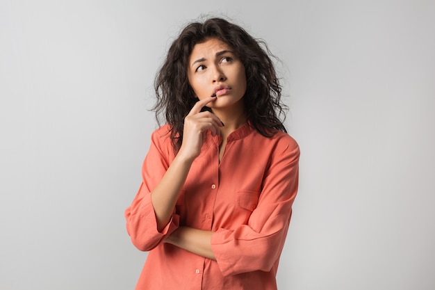 Hübsche junge Frau, die über Problem, verwirrte Emotion nachdenkt, isoliert, orange Hemd, Hipster-Stil, lockiges Haar tragend, Finger an den Lippen haltend, aufblickend, Idee habend