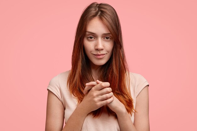 Hübsche junge Frau mit angenehmem Aussehen, hält Hände zusammen und schaut geheimnisvoll in die Kamera, isoliert auf rosa Hintergrund.