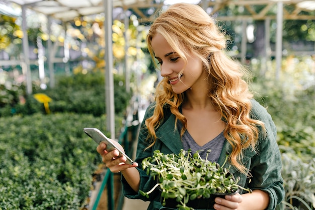 Hübsche junge Frau mit Lächeln liest Nachricht im Telefon. Porträt des Mädchens, das im botanischen Garten im grünen Baumwolloberteil geht.