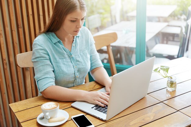 Hübsche junge Frau mit langen Haaren, die im Café mit Laptop sitzen
