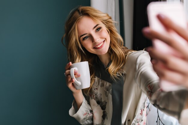 Hübsche junge Frau mit welligem blondem Haar, das selfie nimmt, das neben dem Fenster mit Tasse Kaffee des Morgens, Tee sitzt. Sie trägt einen Seidenpyjama. Türkisfarbene Wand.