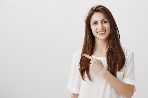 Hübsche junge Frau zeigt mit dem Finger nach links und lächelt als einladender Blick auf den Copyspace