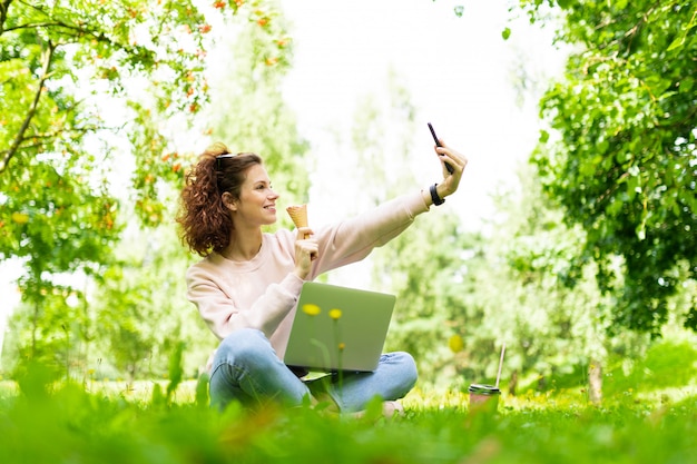 Hübsche junge kaukasische frau ist freiberuflich im park mit laptop, tasse kaffee und eis beschäftigt, sitzt auf einem rasen und macht selfie